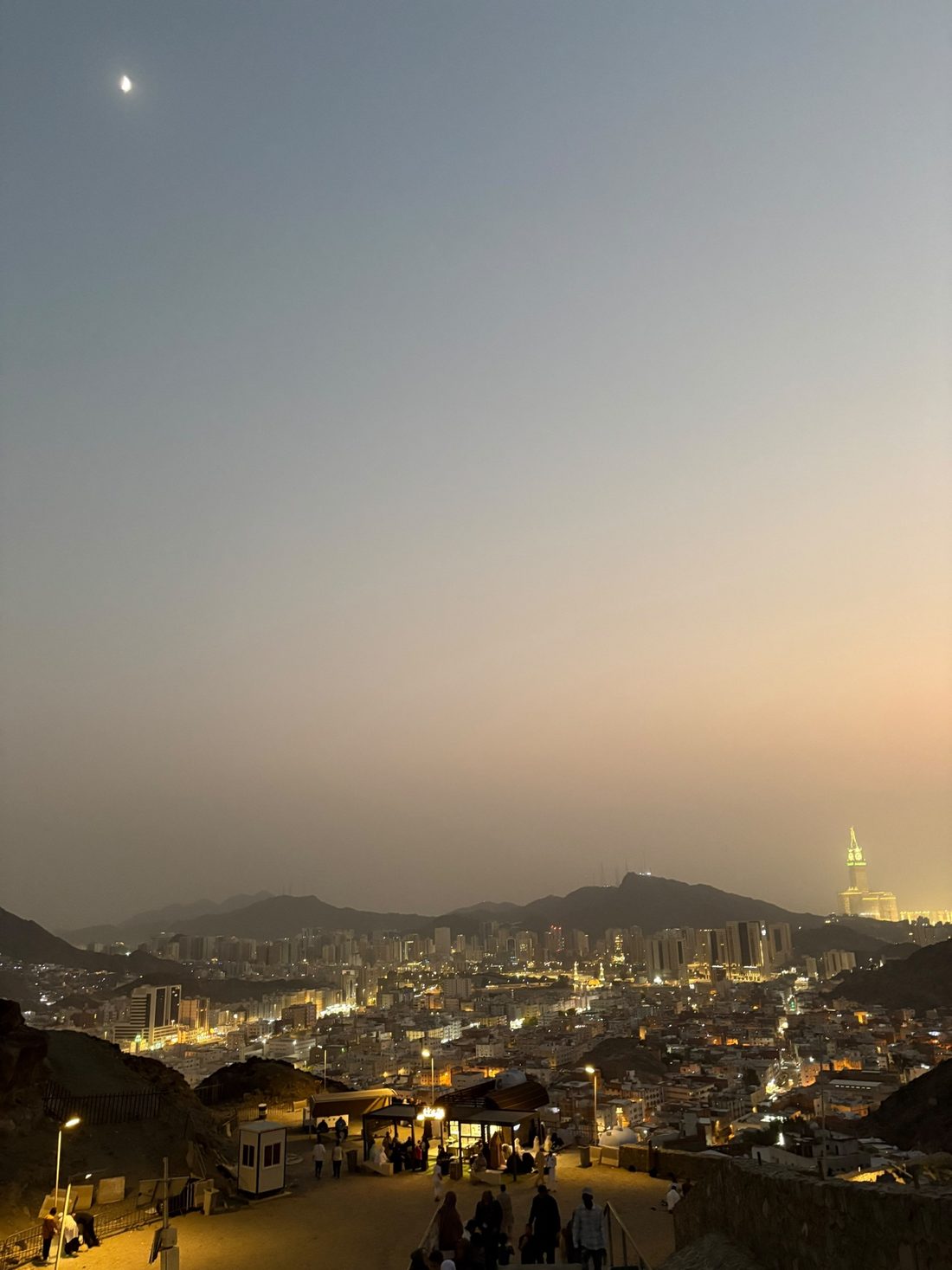 Makkah at dusk seen from the mountain, the clock tower glowing on the horizon under a crescent moon